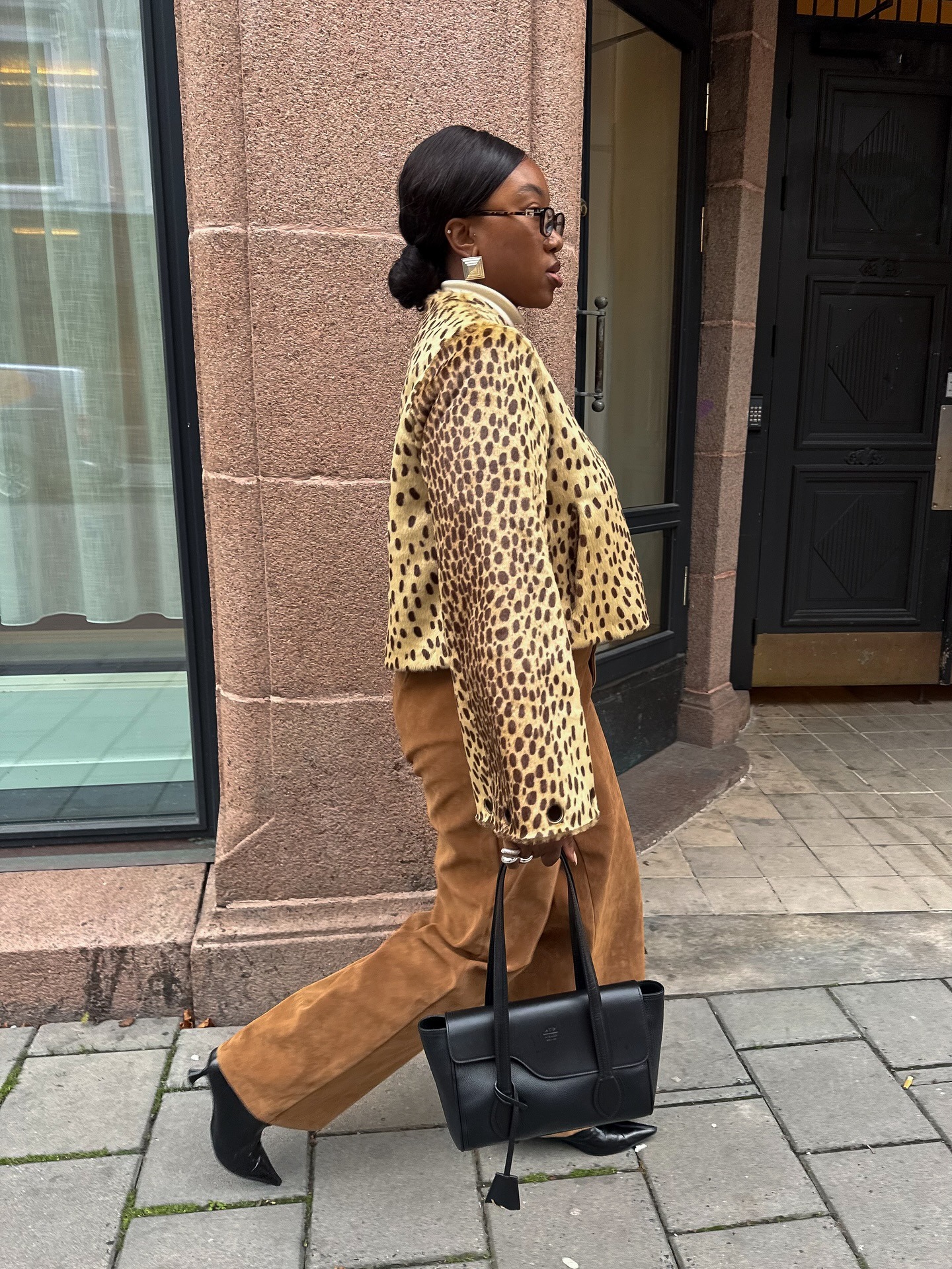 Nnenna holding a black structured bag against brown pants and a cheetah coat.