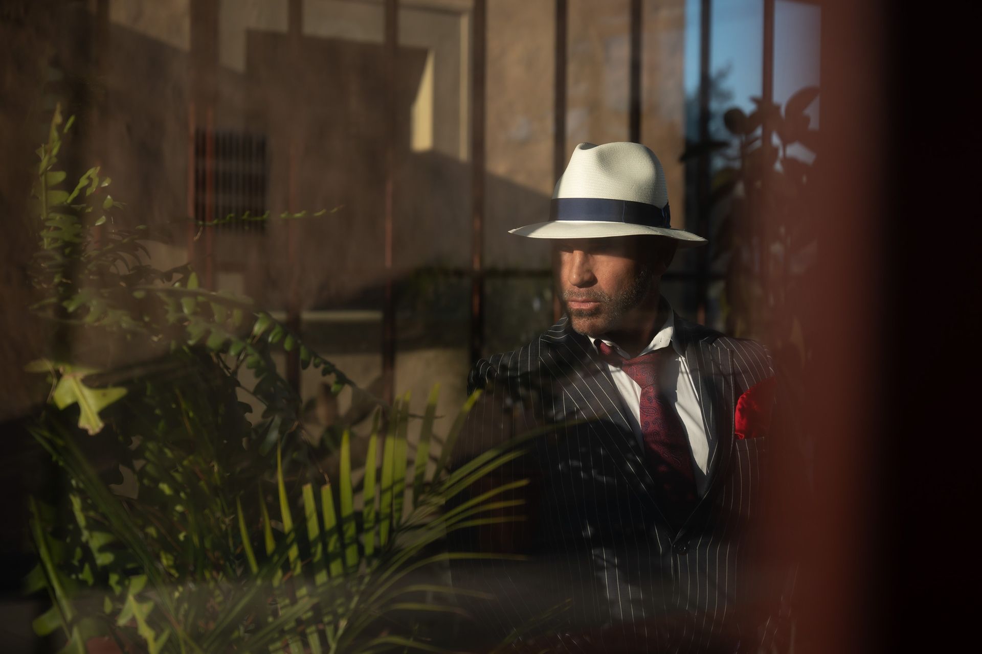 joe gorga wearing a hat and striped suit hiding behind a plant in a widow's reflection in a promo shot for the mob