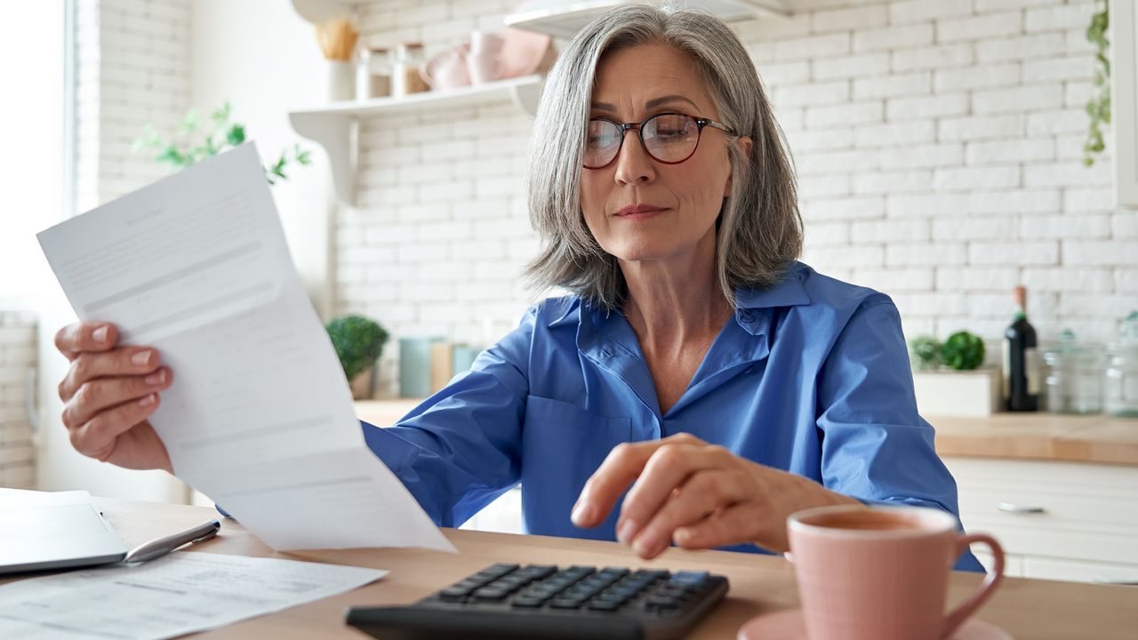 An older woman looks at paperwork and uses a calculator at her kitchen island.