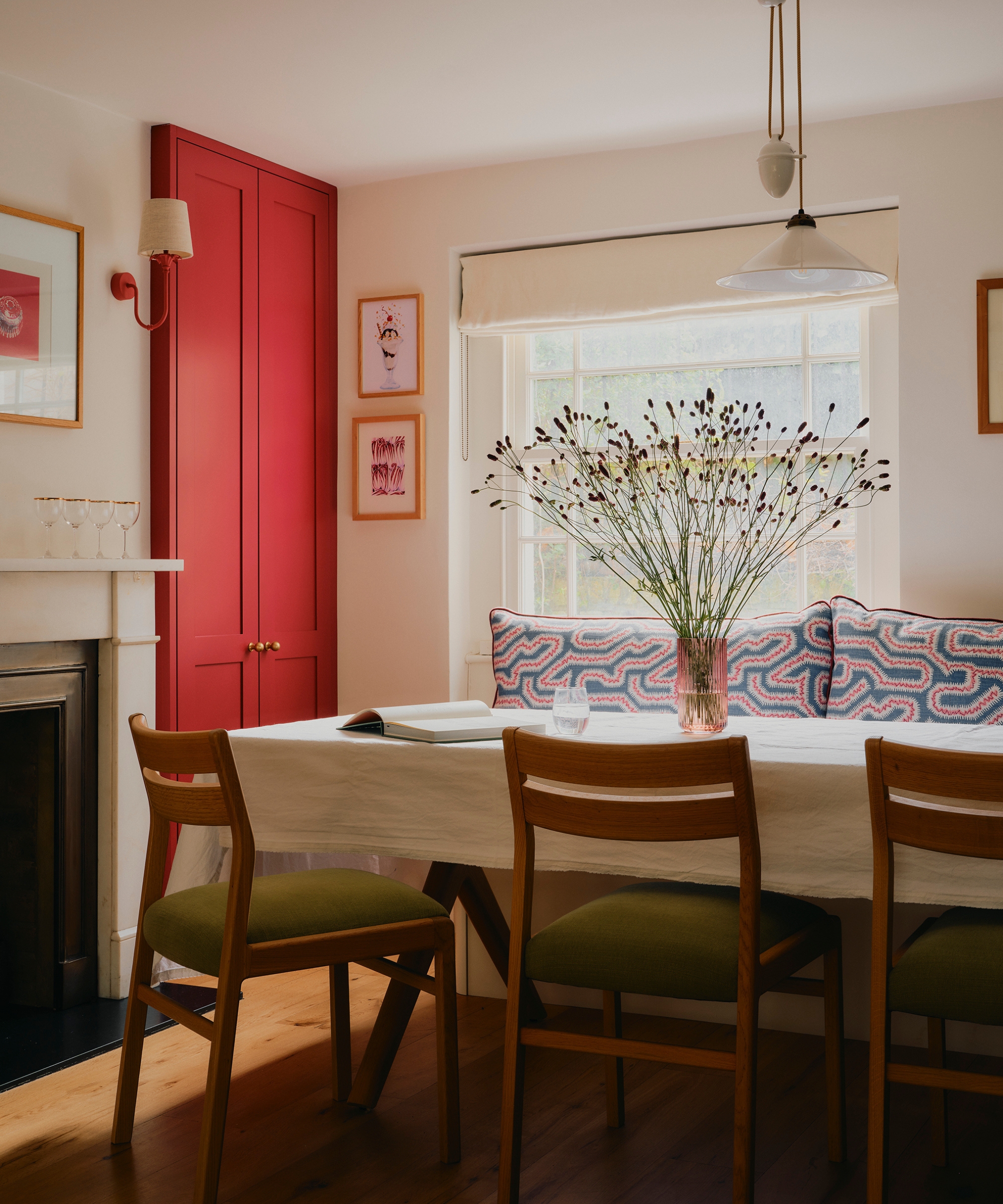 Dining room with a tomato-red built-in cabinet, cream walls, oak dining table, patterned banquette, and mid-century wooden chairs.