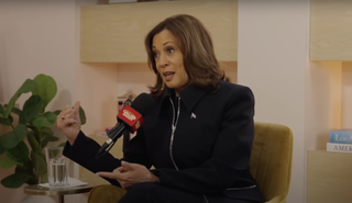 Kamala Harris wearing a black suit and talking into a microphone on the Call Her Daddy podcast