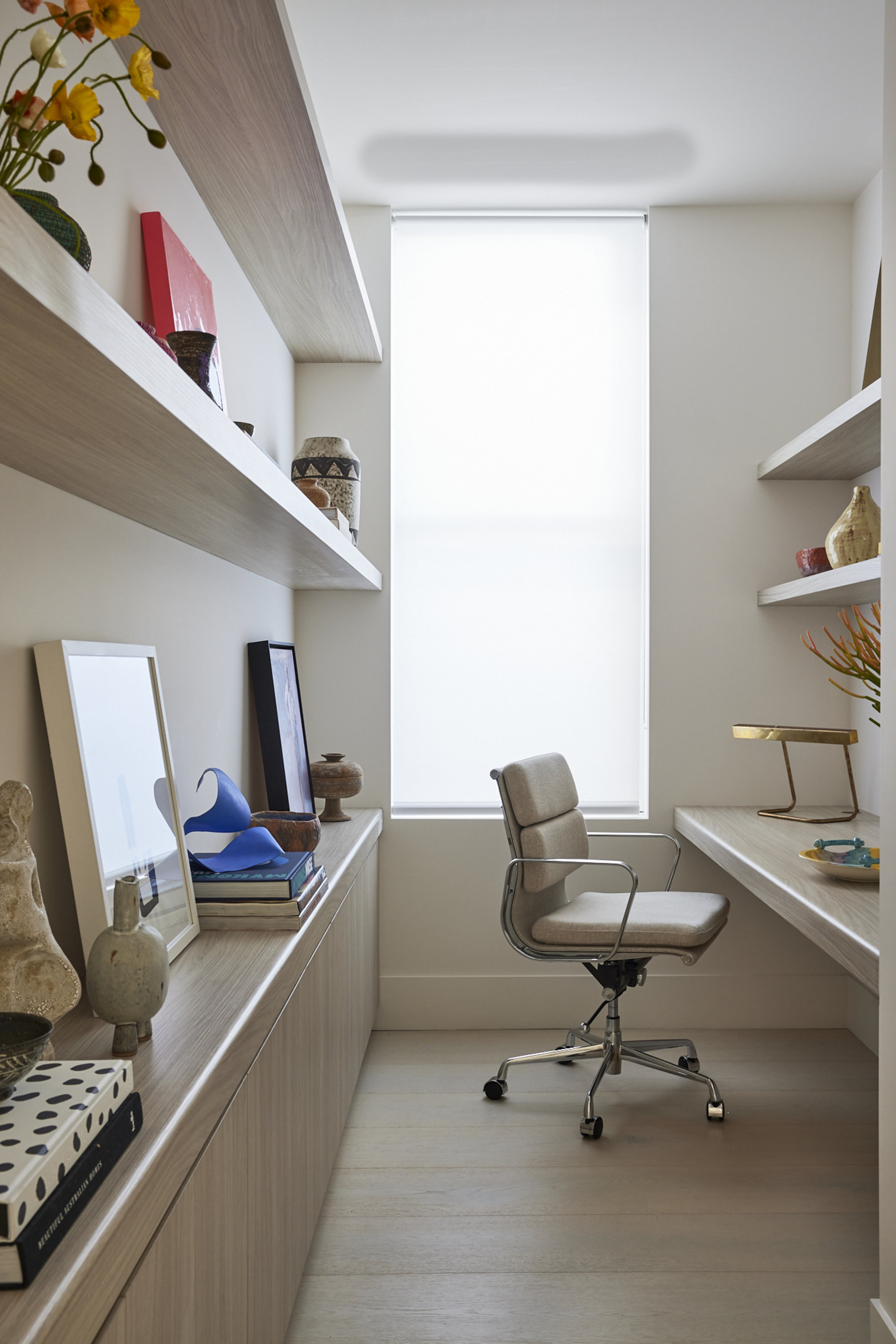 narrow home office with built-in deck and beige desk chair, floating shelves styled with leaning artworks, sculptures and flowers and a window at the end of the room