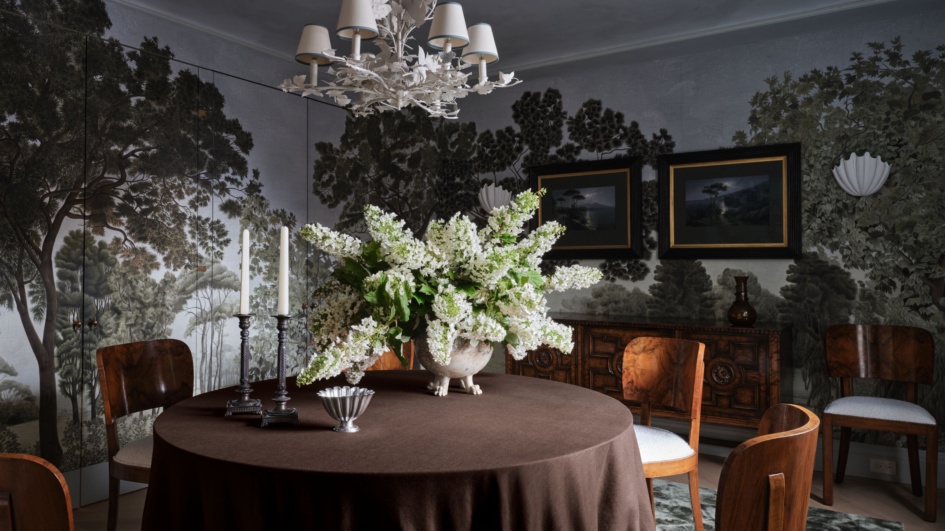 a moody blue grey dining room wrapped in a forest mural wallpaper with round table, vintage wooden chairs and credenza and shell wall sconces