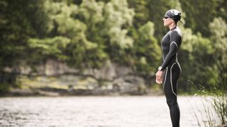 Woman waiting to swim by a river bank in a wetsuit, cap and goggles
