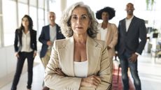 A wealthy older woman with her arms crossed stands with her back to several advisers behind her.