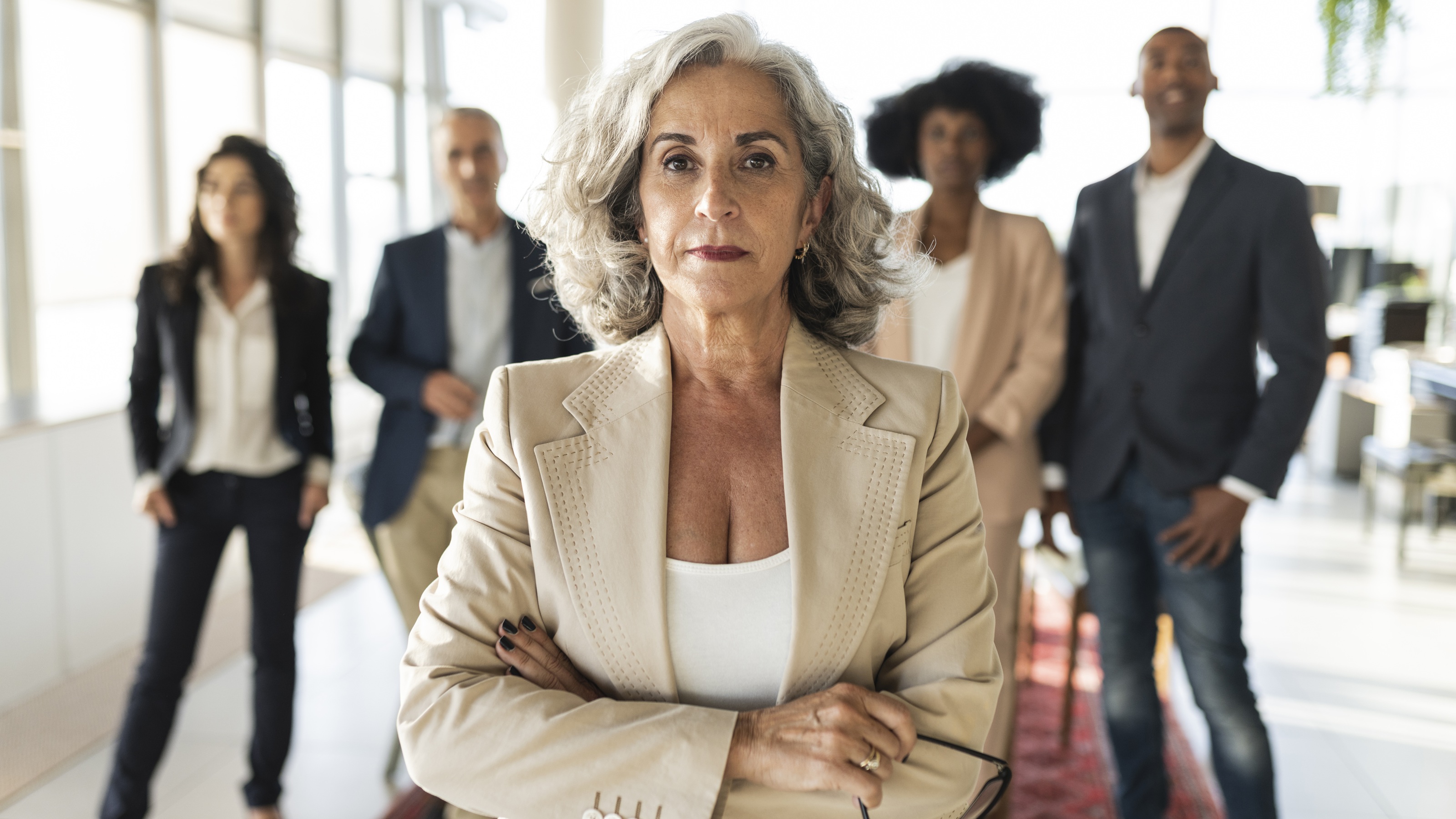 A wealthy older woman with her arms crossed stands with her back to several advisers behind her.