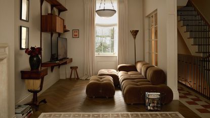 beige living room with khaki sofa, parquetry floors, wall bracket with timber, beige curtains, rug, and timber side table with artwork above, and view of stairs