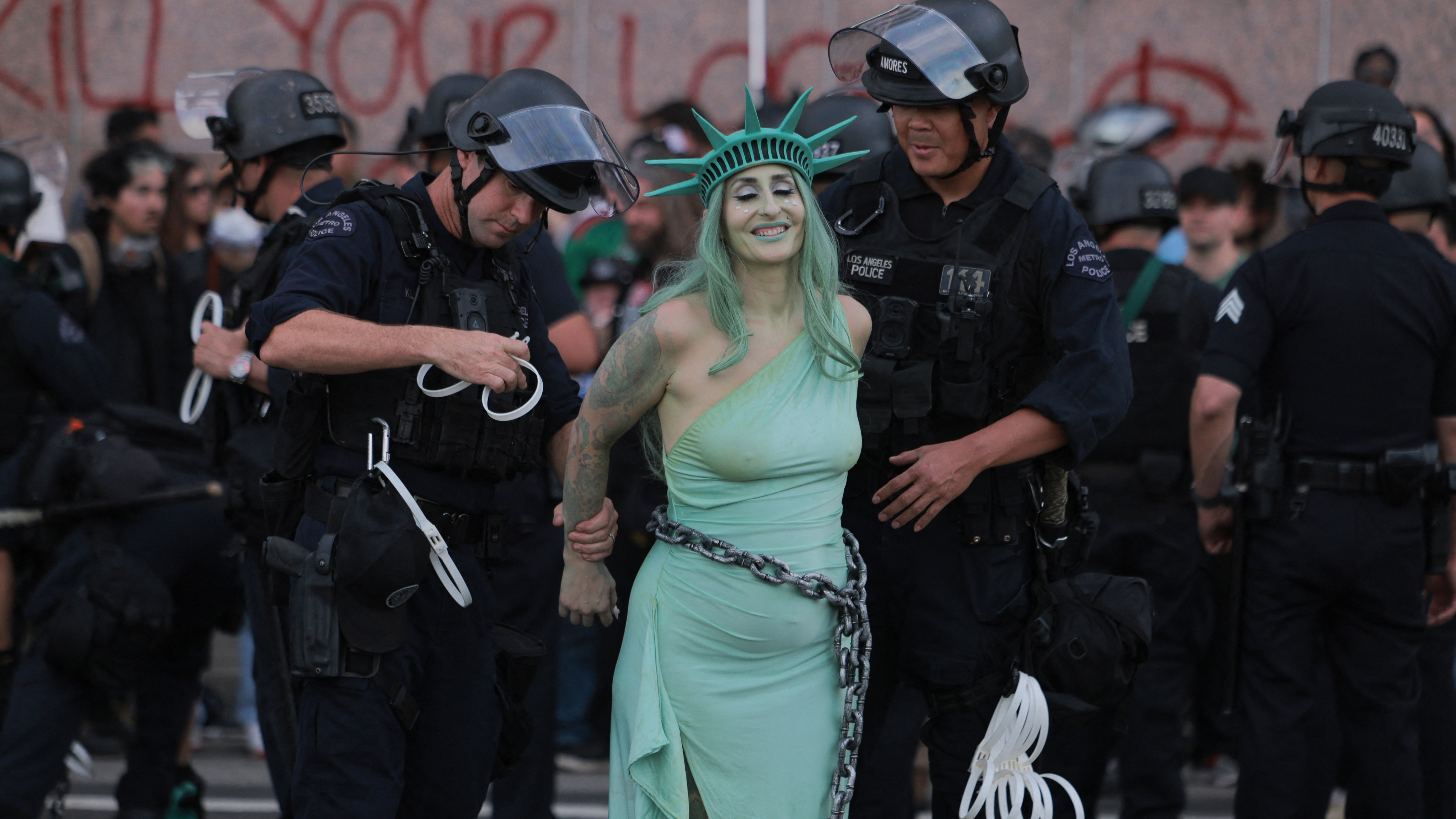 TOPSHOT - LAPD officers arrest a protester dressed as Lady Liberty in chains following clashes near the Metropolitan Detention Center during the "No Kings" national day of protest in Los Angeles on March 28, 2026. Huge crowds of protesters rallied across the United States on March 28 against President Donald Trump, venting their fury over what they see as his authoritarian style of governing, his hardline immigration policies and the war with Iran. Organizers said "at least 8 million people gathered today at more than 3,300 events across all 50 states," from big cities and small towns. US authorities provided no national crowd estimate. (Photo by ETIENNE LAURENT / AFP via Getty Images)