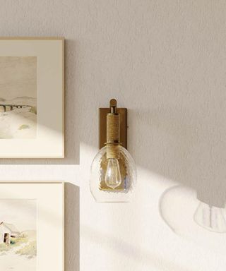 A brass and glass wall light on a white wall next to two framed landscape illustrations hung vertically