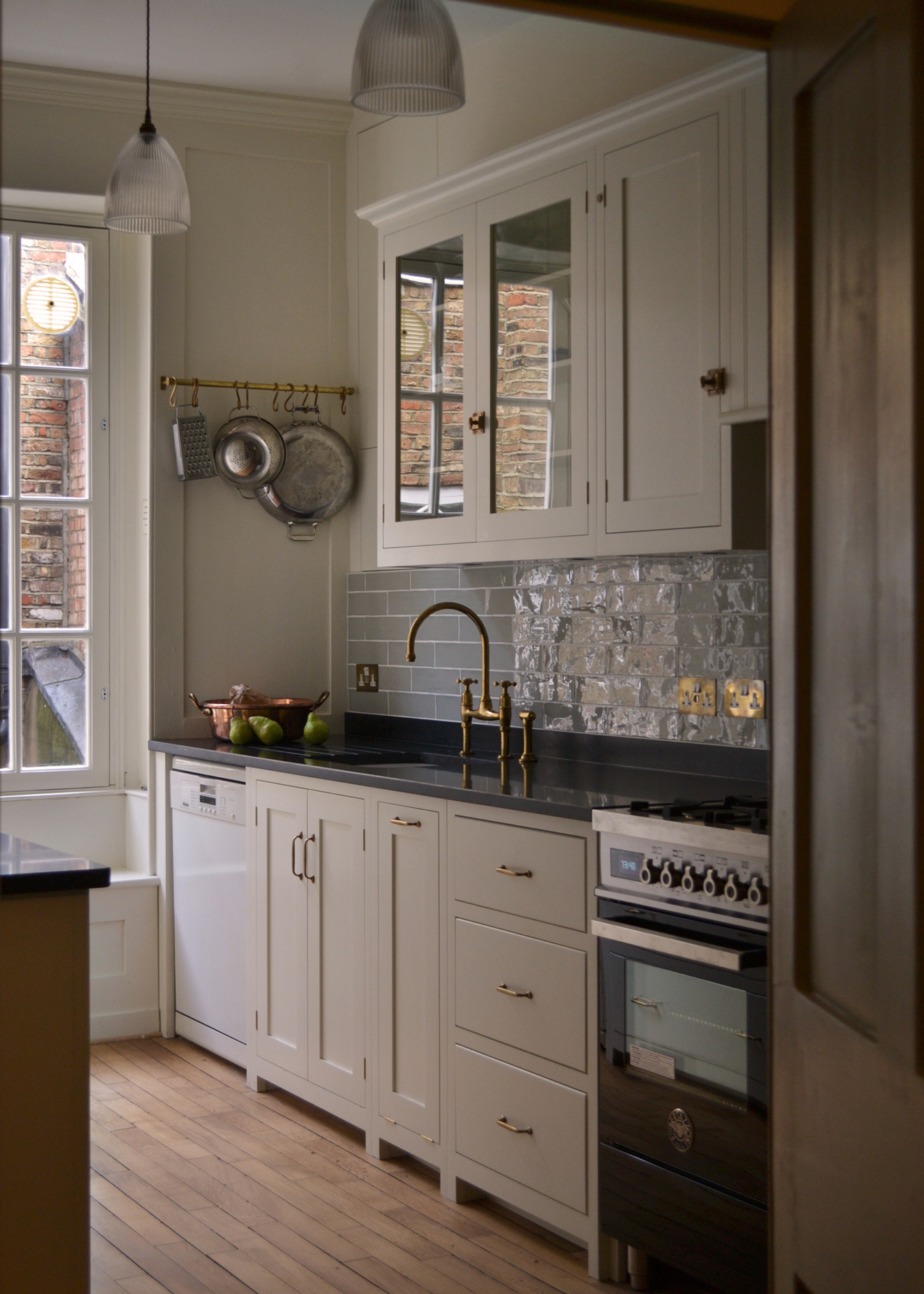 a small narrow kitchen with creamy cabinets and subway tiles. In one corner is a brass rail for pans