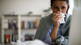 A female employee blowing her nose while wrapped in a blanket and looking at her laptop, to represent Blue Monday. The background is a blurred office.