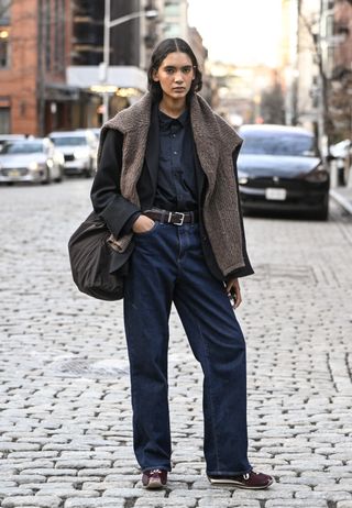 A woman wears a denim top, jeans, loewe runners, and a brown suede around her shoulders.