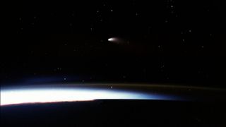 a faint comet with bright short tail hangs in space over a dark curve of earth, slightly illuminated in the atmosphere.