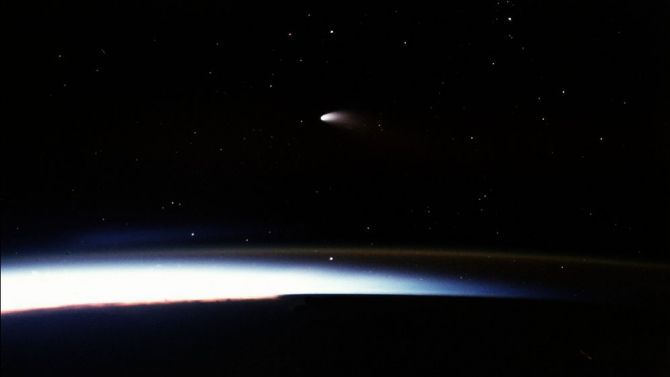 a faint comet with bright short tail hangs in space over a dark curve of earth, slightly illuminated in the atmosphere.