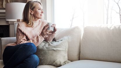 An older woman looks out the window while sitting on her sofa with a cup of coffee.