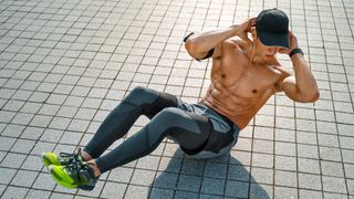 Muscular man doing twist abs exercise with raised legs. Photo of young man workout along outdoor in the city