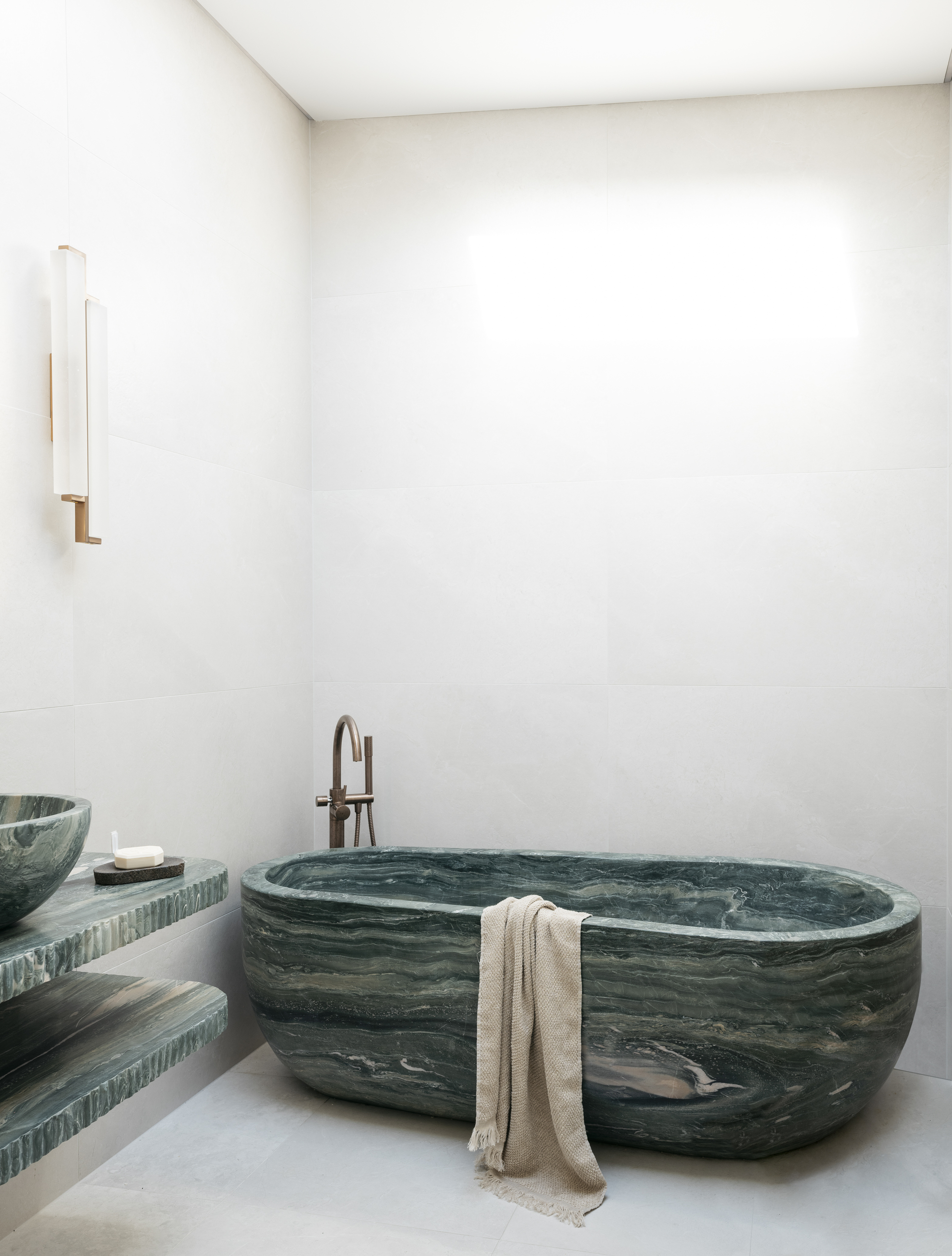 neutral bathroom with green marble freestanding bath, floating green marble vanity, wall sconce, and towel draped over the edge of the bath