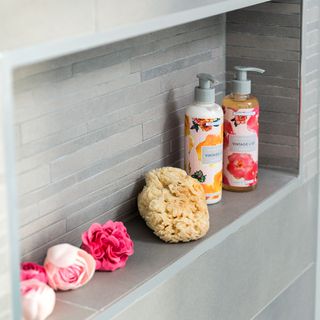 A recessed shelf in a shower with bottles, sponge and flowers on