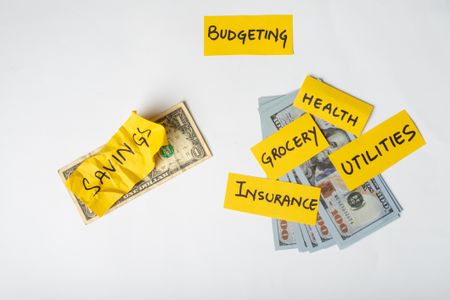 a crumpled yellow sticky note labeled "savings" placed on a single US dollar bill, in contrast with a pile of hundred-dollar bills with other yellow sticky notes for significant household expenses
