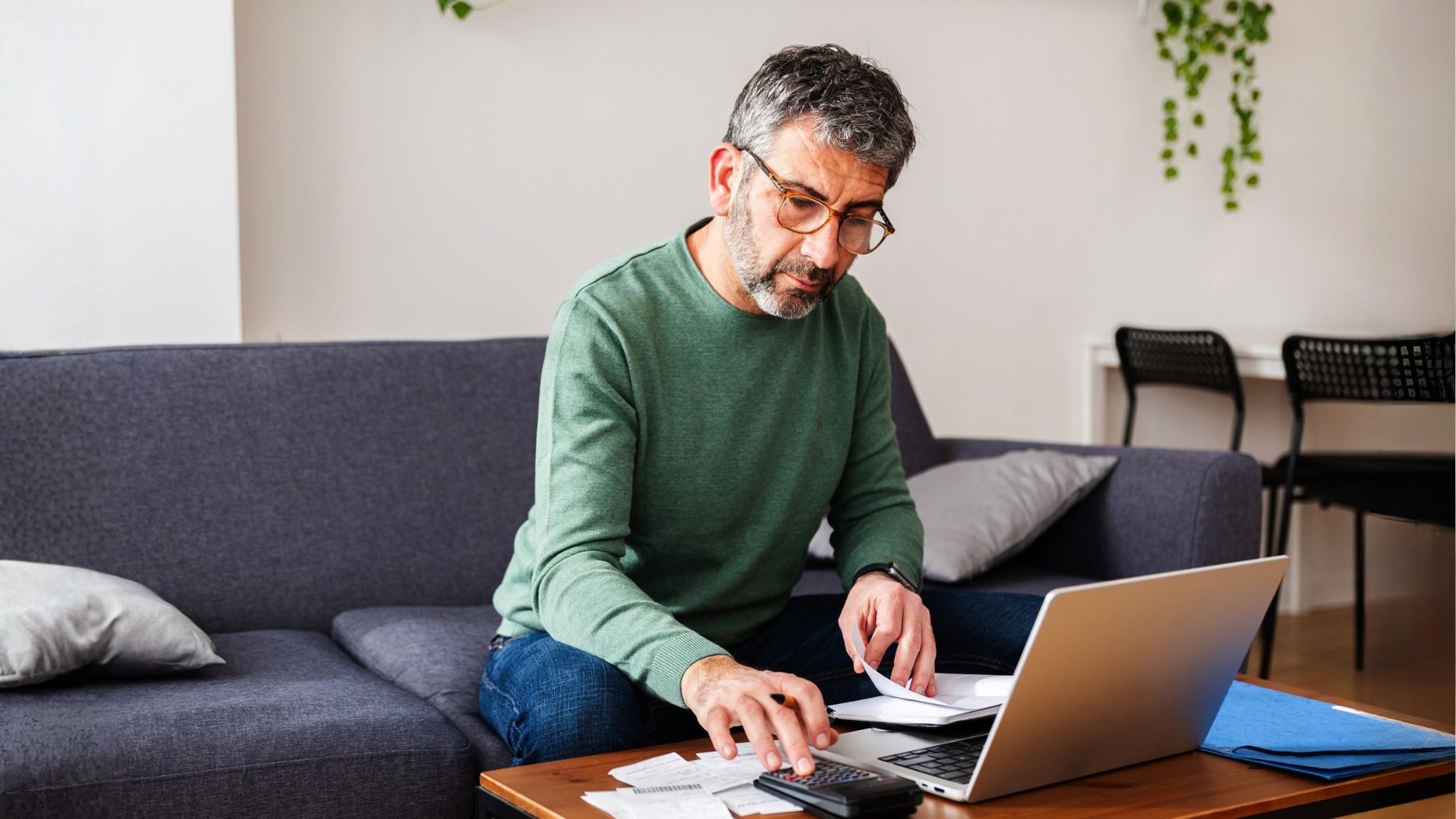Middle aged man calculating expenses and managing the budget while using a calculator and laptop at home