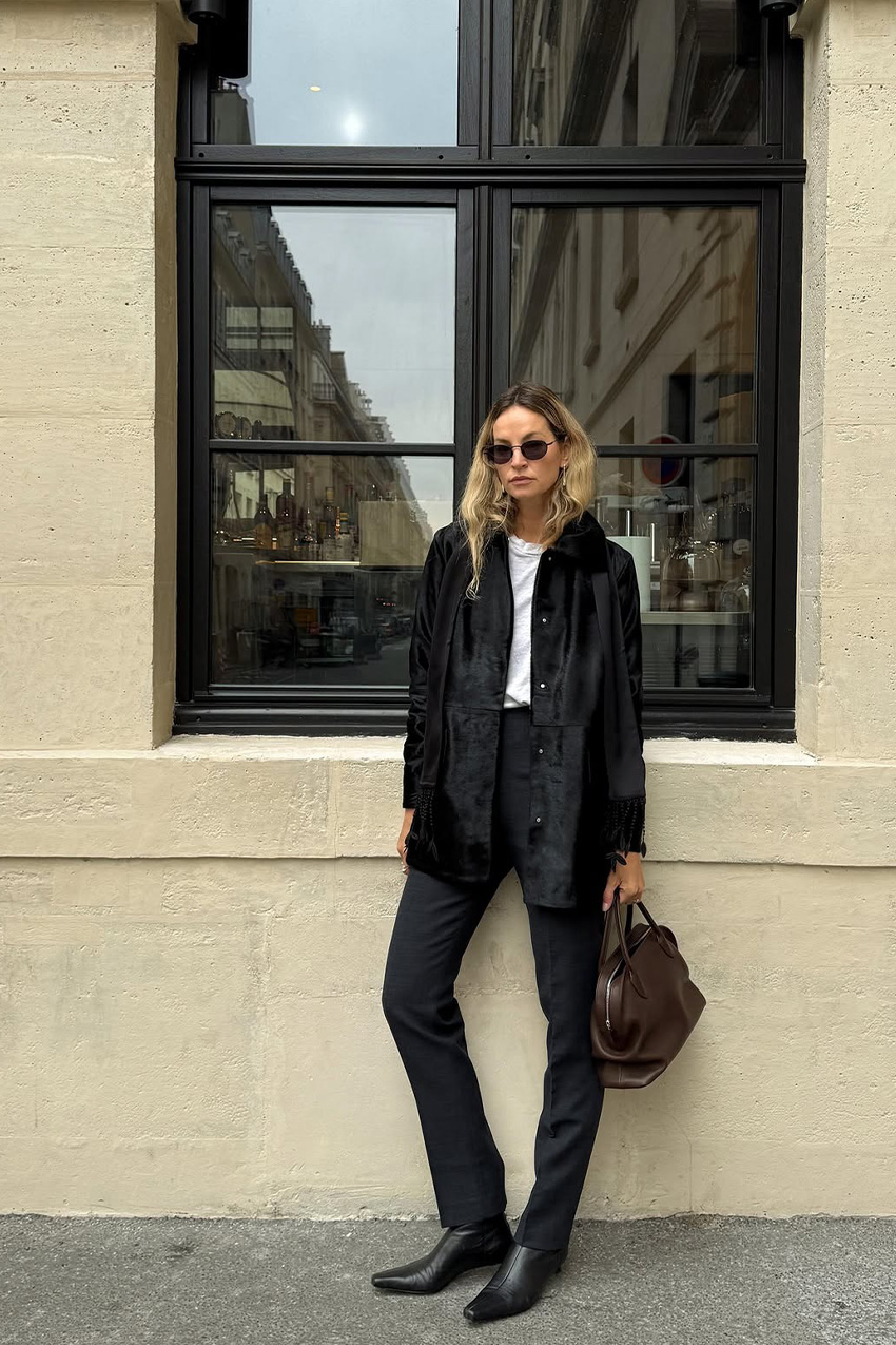 How to wear skinny jeans is shown in a photo of a woman leaning against a building outside, wearing oversized aviators with a black pony hair coat styled over a white t-shirt paired with black cigarette-style jeans, a brown bowler necklace, and black pointed ankle boots