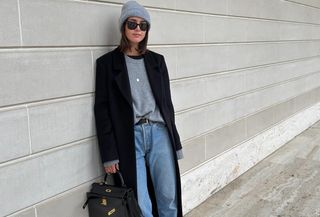 A woman wears a gray hat, gray sweater, black coat, black belt, Hermes bag, and jeans.