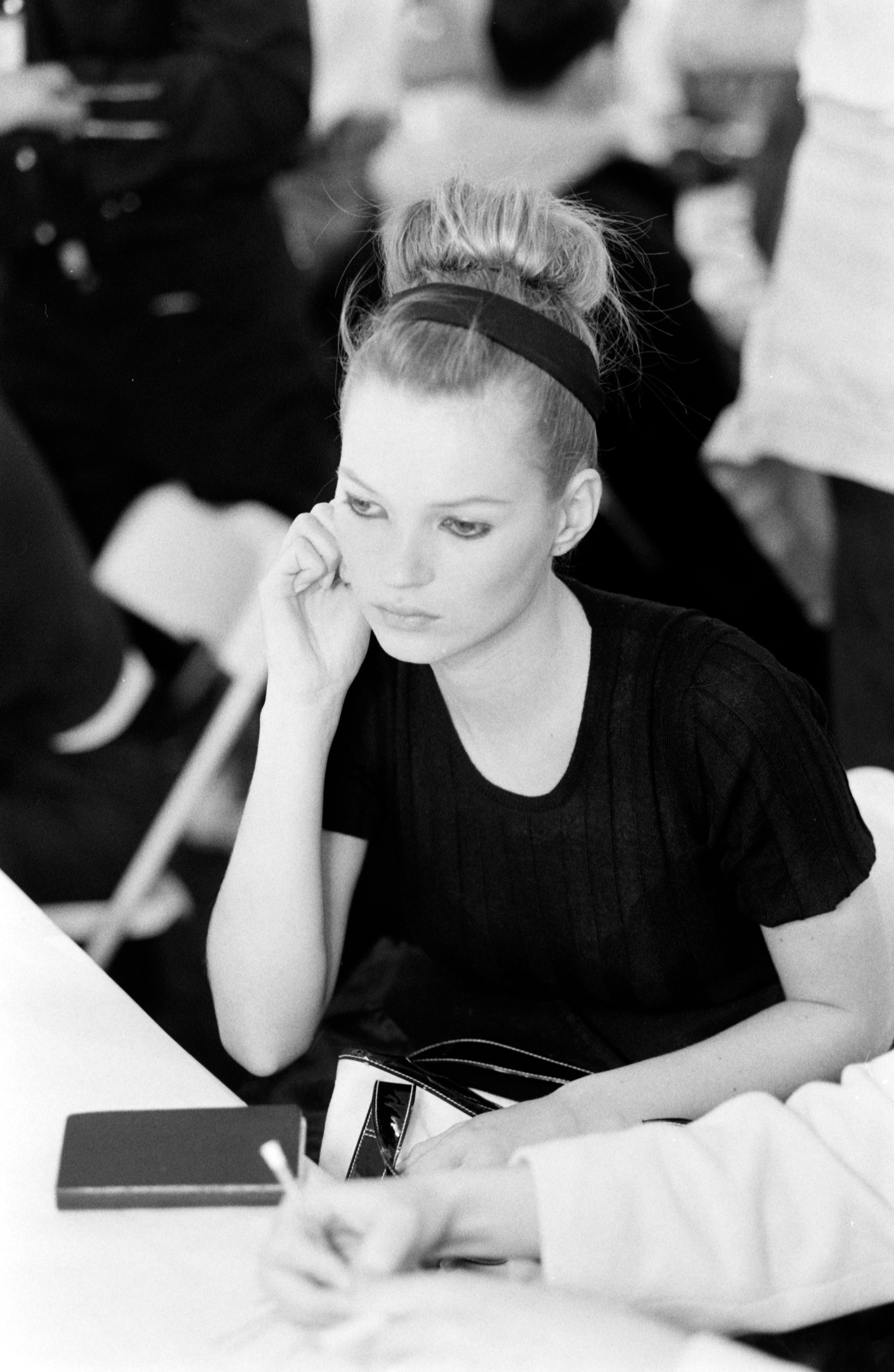 Black-and-white photograph of Kate Moss sitting backstage at a fashion show, resting her face against one hand and looking pensively to the side, wearing a black top with her hair in a high bun.