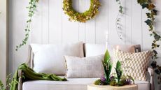 Bright white sunroom featuring a white sofa accented with a green throw blanket and patterned throw pillows. Elsewhere, there is a stone side table holding lily of the valley taper holders and a shamrock wreath hanging overhead