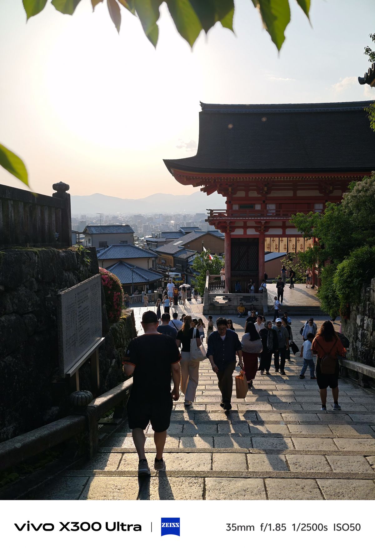 People walking down a stone staircase next to traditional temple architecture during a warm, hazy sunset over the city.