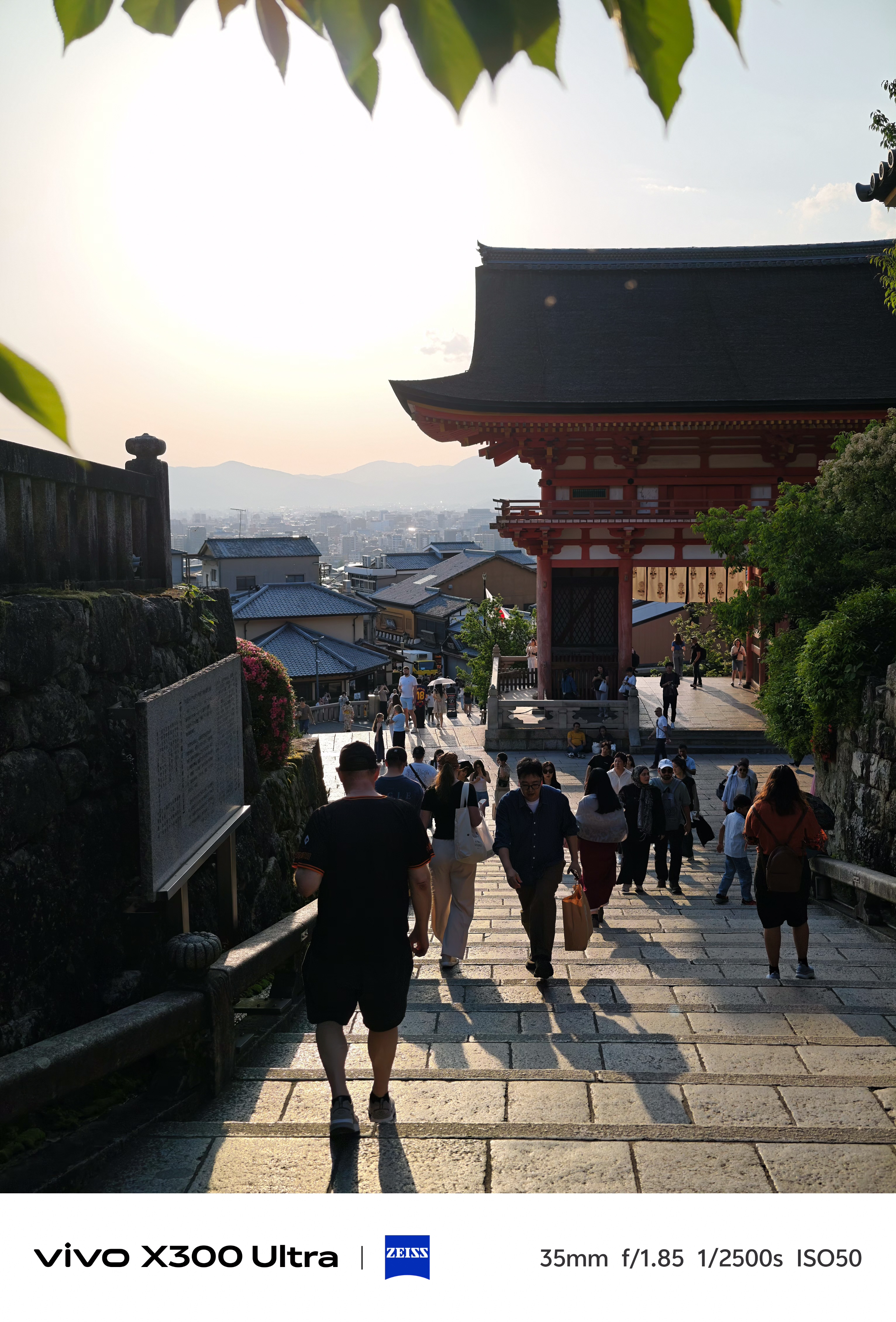 People walking down a stone staircase next to traditional temple architecture during a warm, hazy sunset over the city.