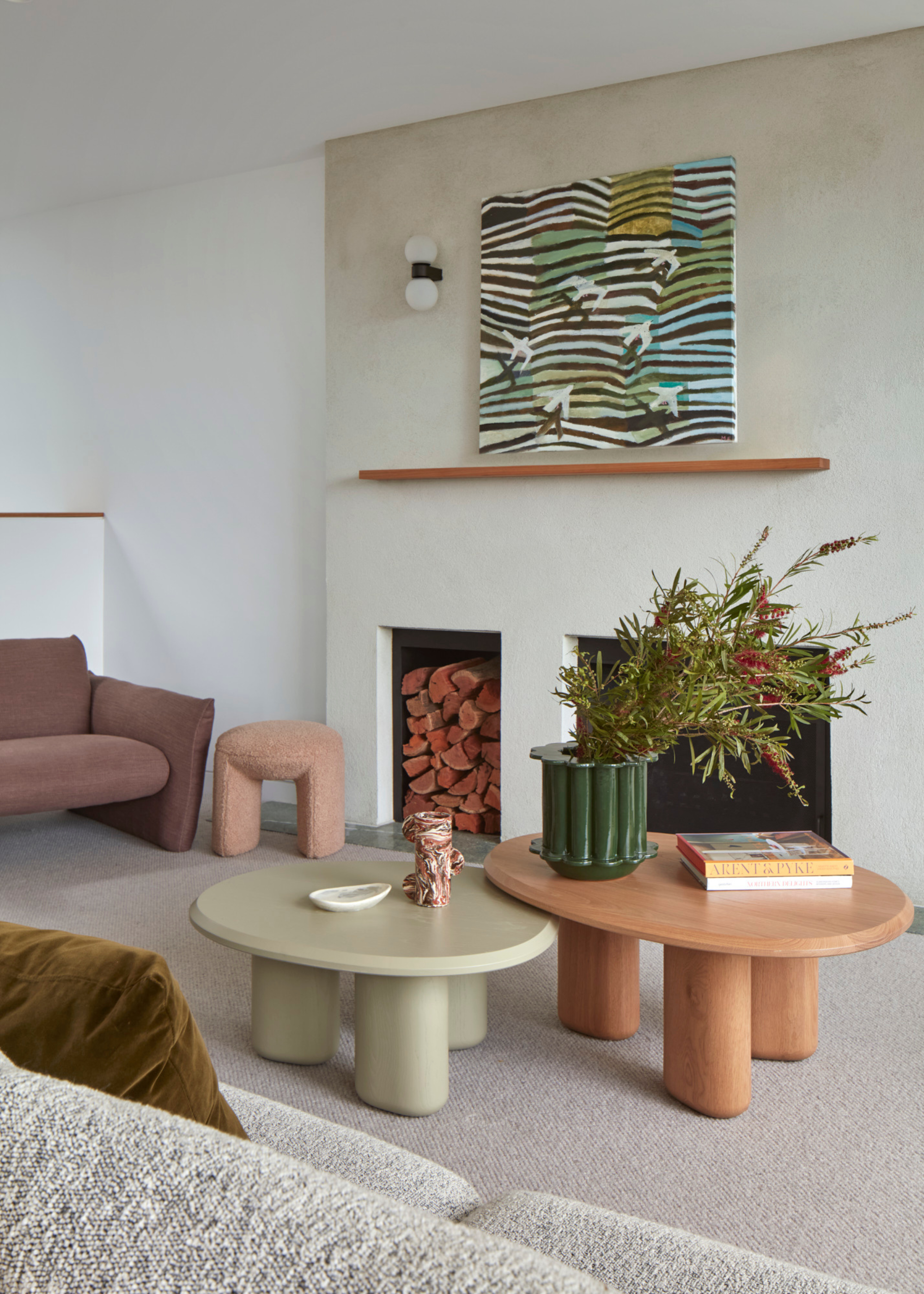 A living room with a wood and a sage coffee table set, a stack of books, a vase with stems, a floating mantel above a fireplace with hanging art, and a blush pouf below