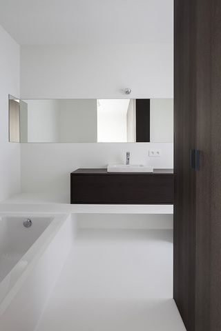 The bathroom is all white, with dark wood details. The bathtub is to the left and the sink area to the far wall, with a mirror above it.