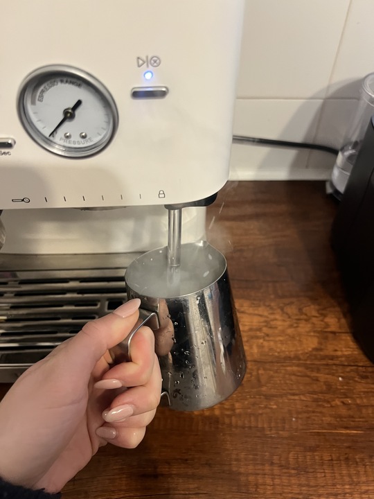Image of a hand holding a milk jug under a steaming wand attached to a white espresso machine over a wooden countertop. There is steam coming out of the jug.
