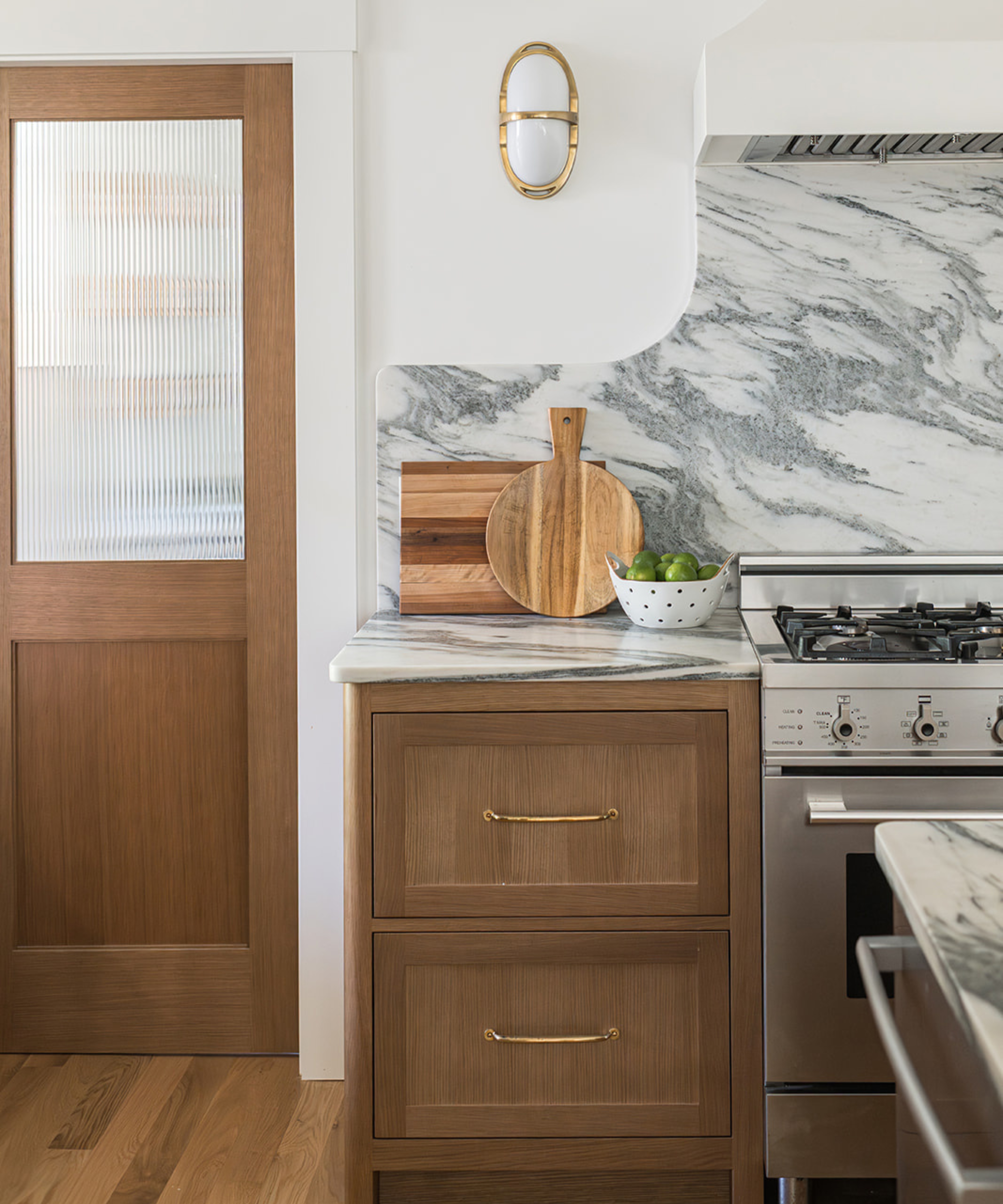 A close-up of a kitchen corner featuring natural wood cabinetry with thin brass handles, a dramatic grey-veined marble backsplash, and a fluted glass door