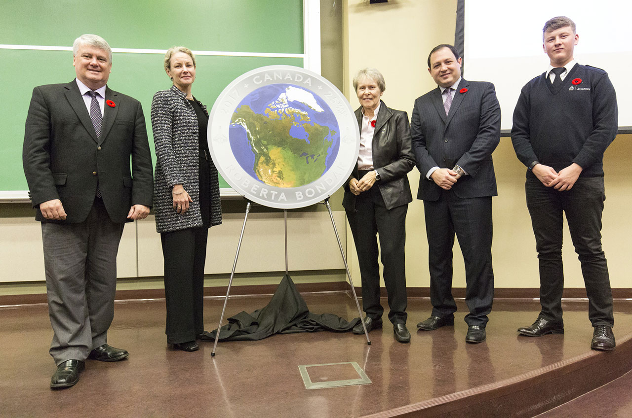 Five people pose with a large model of a colorful coin with a map of Canada on it