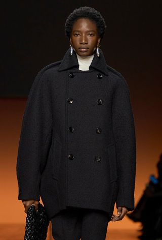 A model from Bottega Veneta's fall 2024 runway wearing a white turtleneck under a black cropped peacoat styled with black pants, a black bag, and silver earrings.