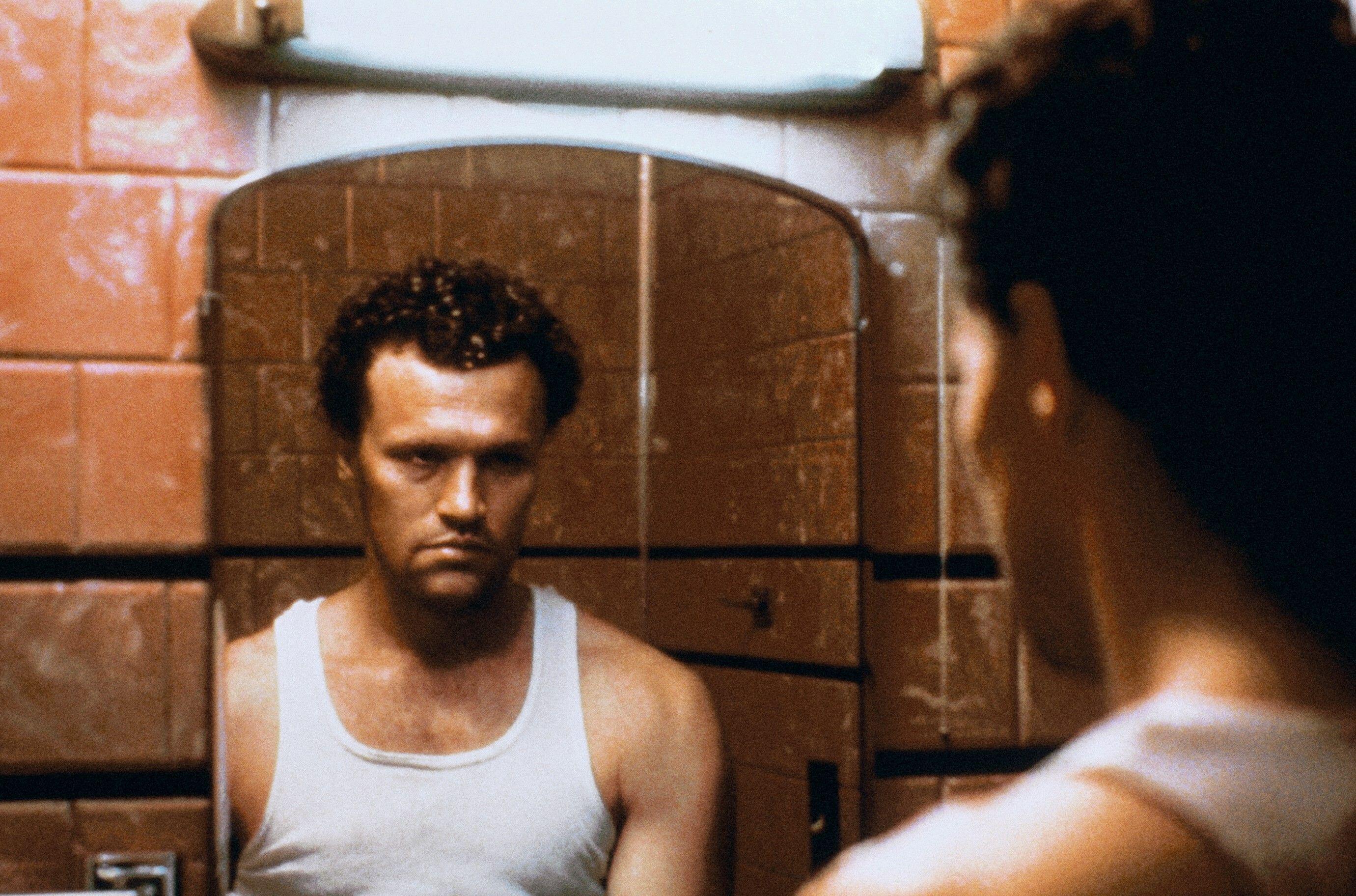 a man in a white tanktop stares in a mirror in the movie henry portrait of a serial killer