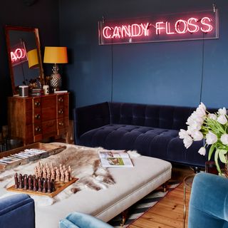 blue living room with a red neon candy floss sign on the wall, dark blue couch with large square cushions table in the middle, with a chess board on top
