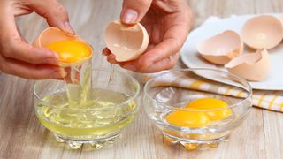 Close up image of a person's hands holding a freshly broken raw egg over a bowl and dumping the egg white into the bowl while keeping the yolk in the broken egg shell. A second bowl with only egg yolks in it sits nearby.