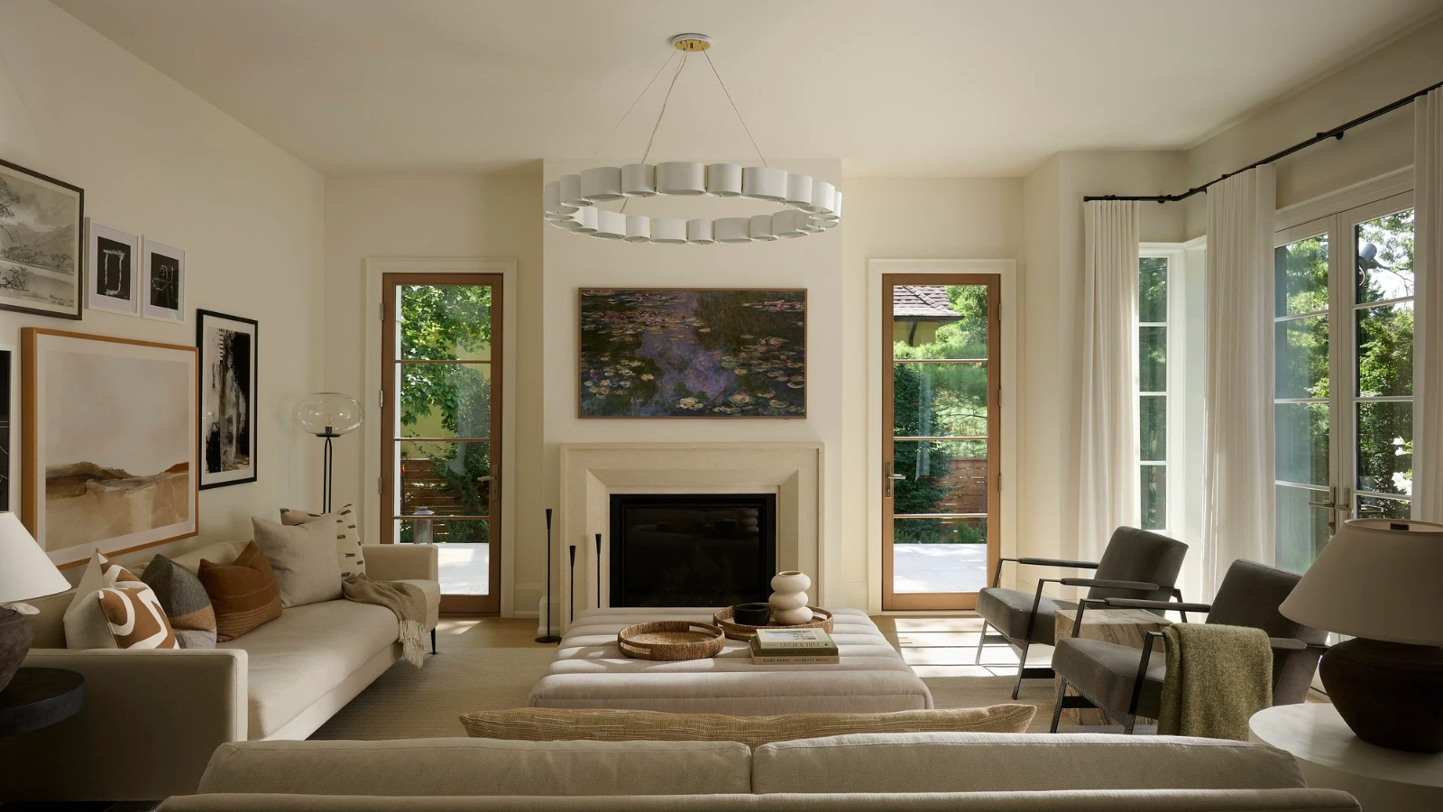 a large living room in a calming, neutral color scheme with artwork hanging above the fireplace, two accent chairs on the right, a large ottoman, two couches, a gallery wall on the left, and a large chandelier in the center of the room