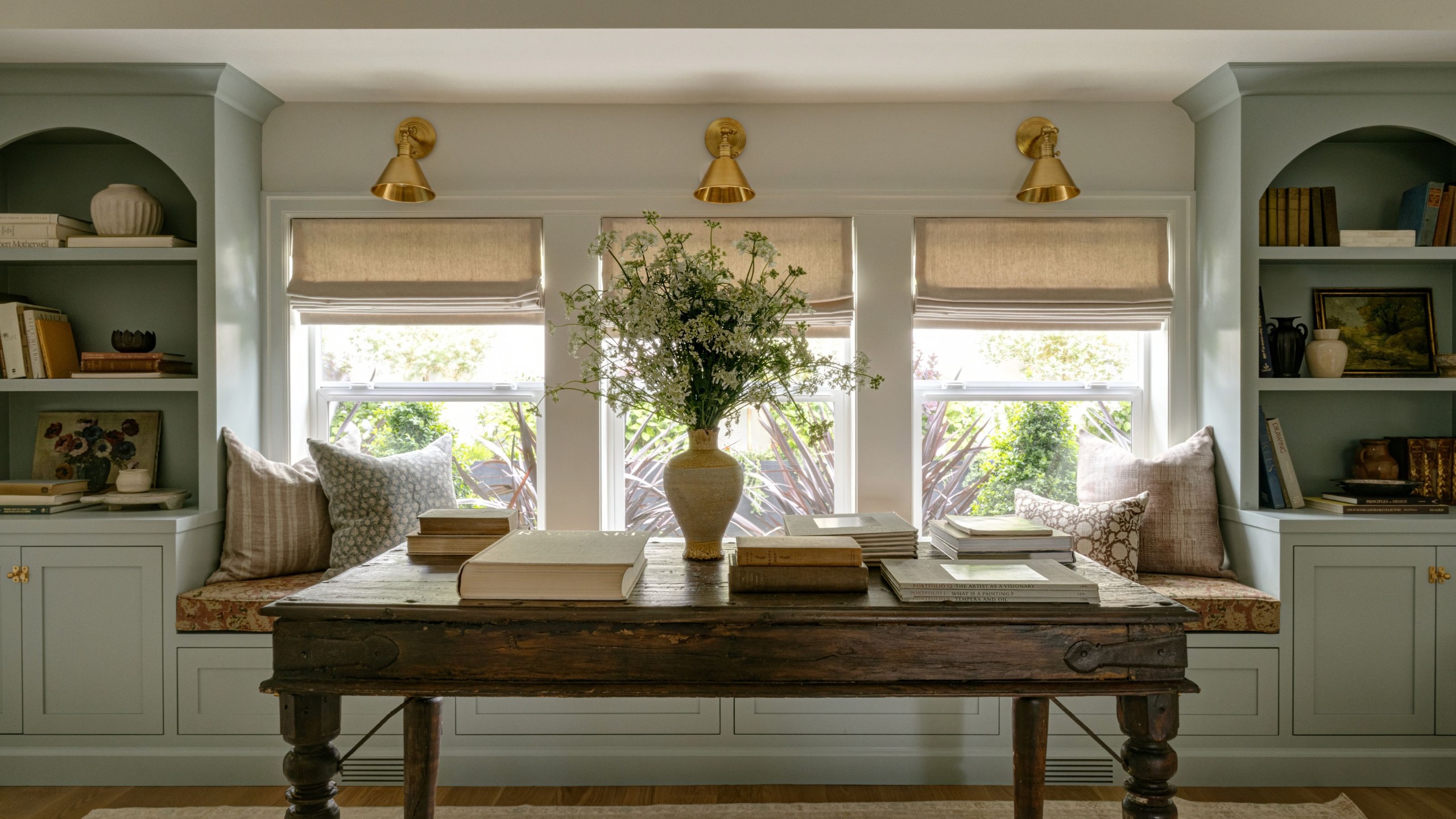 A library room with a wall of built-ins with closed storage and arched open shelving, painted a light gray-green. A window seat in the middle with three windows, neutral Roman blinds, patterned bench upholstery and cushions, and a dark wooden desk.