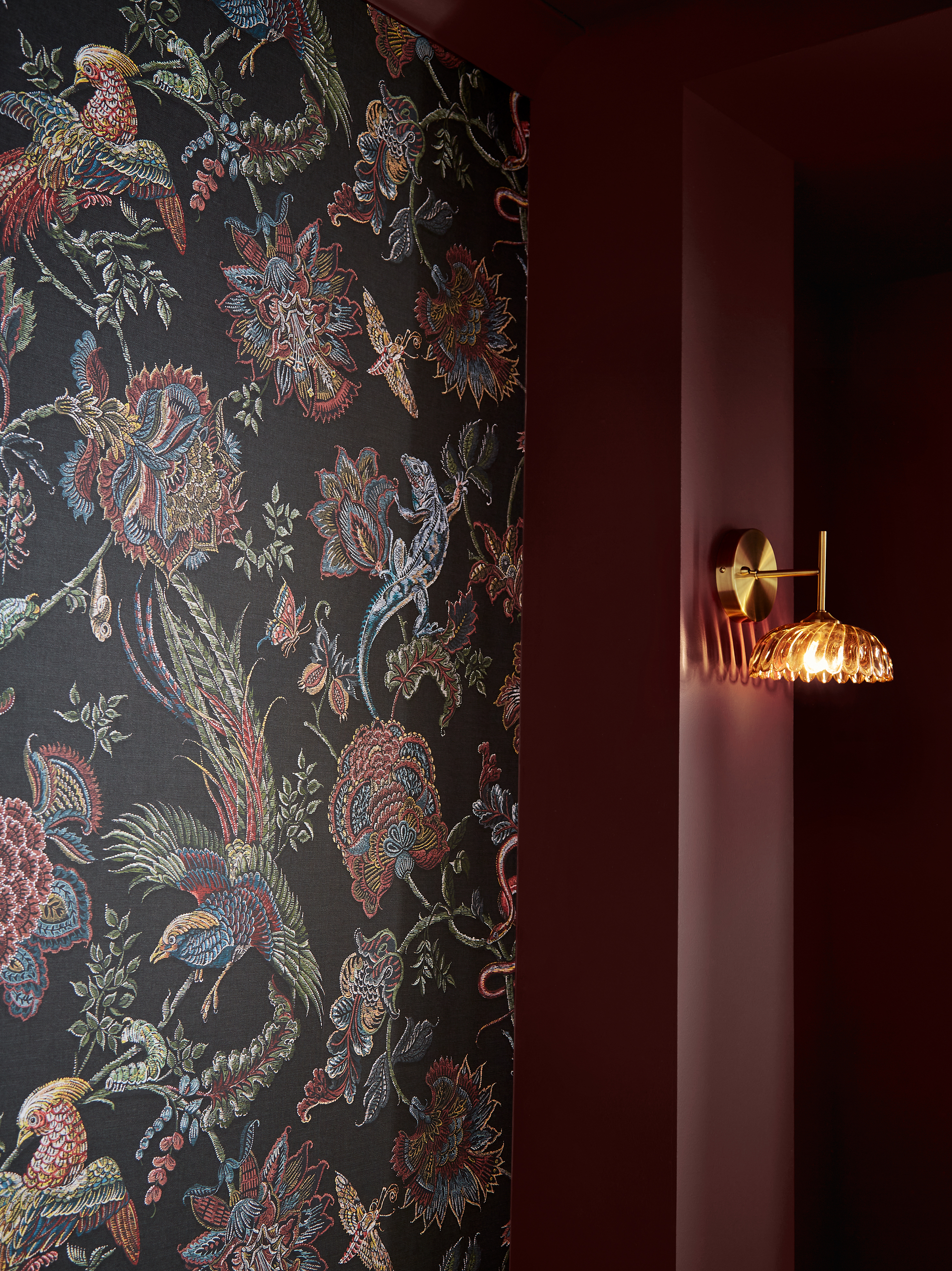 Close-up image of a wall that is covered in a hand-painted wallpaper that is black with multicolored imagery of birds and flowers. The other part of the wall is painted maroon red and there is a gold scone light.