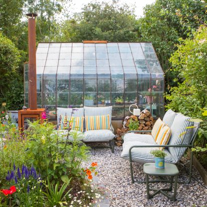 A gravel garden with two John Lewis Marcy 2-seater sofas in green stripe in front of a greenhouse