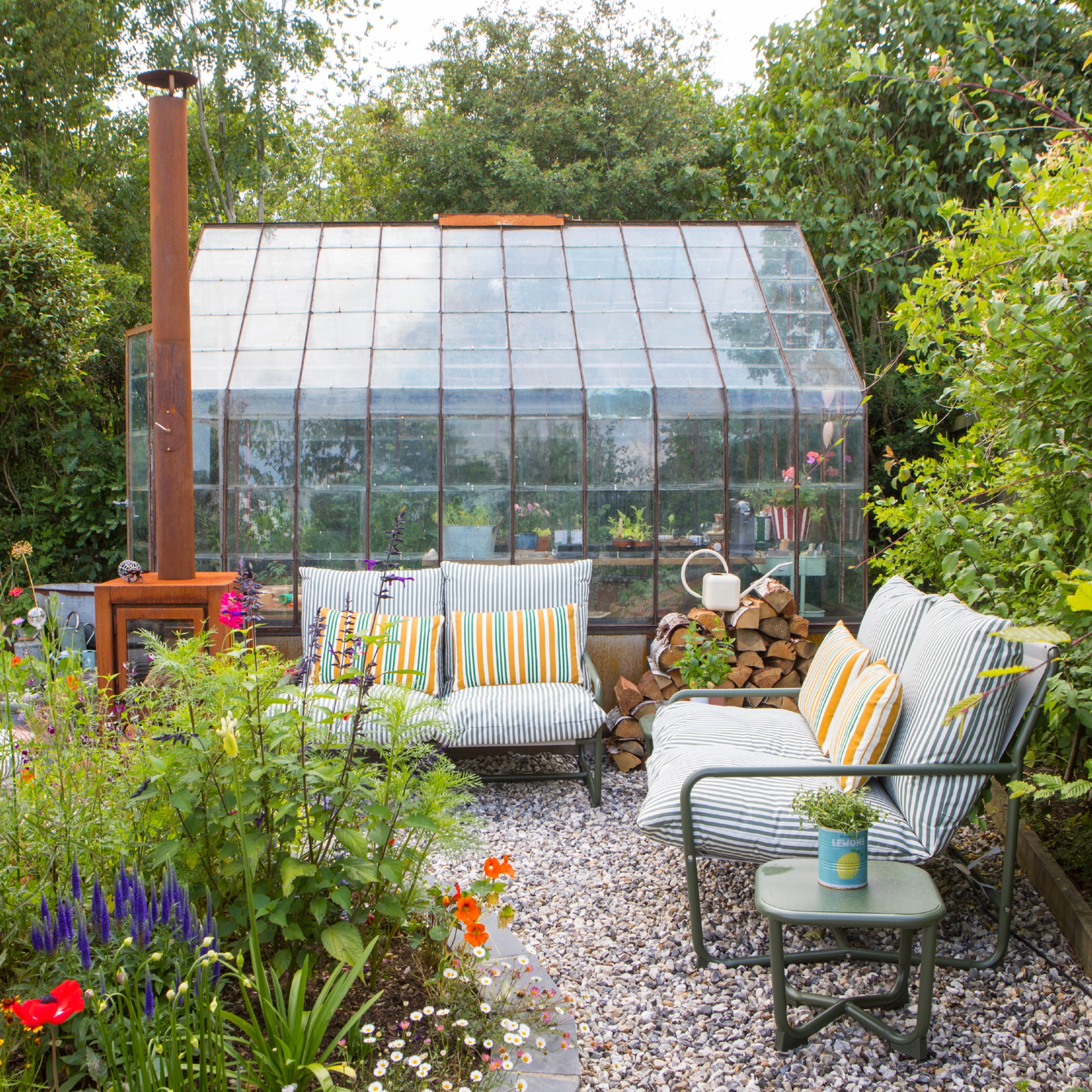 A gravel garden with two John Lewis Marcy 2-seater sofas in green stripe in front of a greenhouse