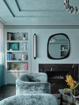 Image of a living room color-drenched in a cyan paint color. The walls and ceiling are painted in cyan as well as the built-in bookcase. There is a black marble fireplace with a black organically-shaped mirror hanging above it. There are two teal, velvet accent chairs in front of the fireplace.