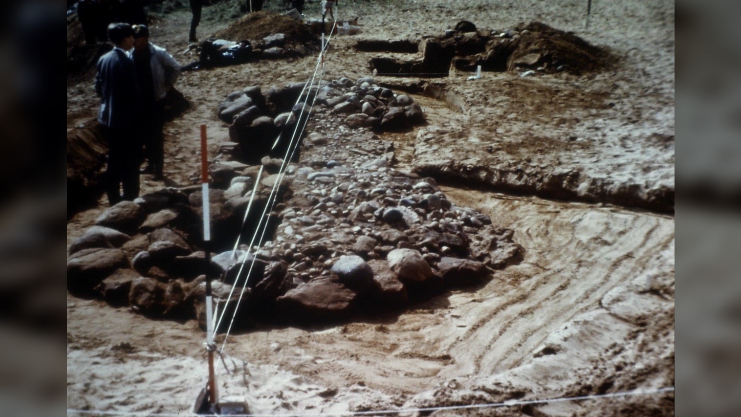 Photograph of the 1965 Lundin Links excavation showing burial up close. Here we see a circle of stones on a beach and two people standing over them.