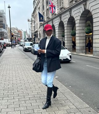 @nlmarilyn wears blue skinny jeans, a white roll-neck jumper, black blazer, a red baseball cap and black knee-high boots