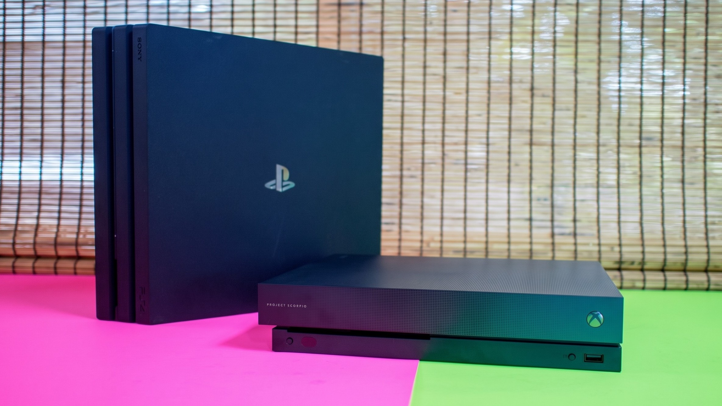 A photo of a black PS4 Pro and a Project Scorpio edition Xbox One X on a pink and green base.