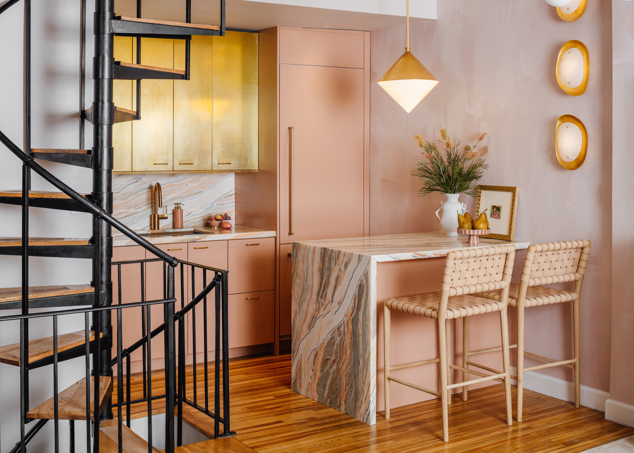 a modern kitchen with brushed gold and blush pink kitchen cabinets with a marble breakfast bar next to a black spiral staircase