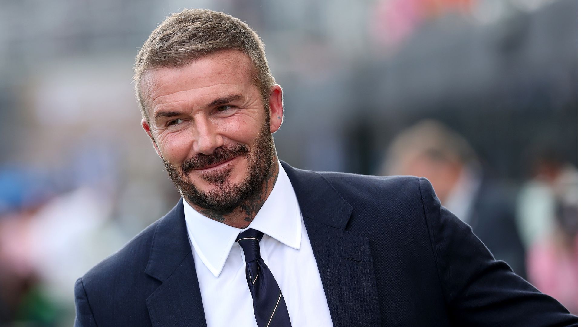 David Beckham smiles, wearing a suit at a football match